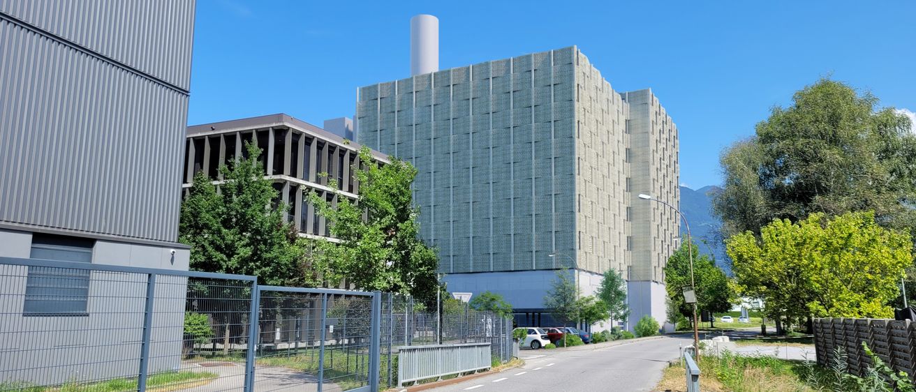 Ein Industriegebäude mit Schornstein steht neben einer Straße, die von einem blauen Tor eingezäunt ist. Bäume und Straßenlaternen säumen die Straße. Ein Auto ist in der Nähe geparkt.