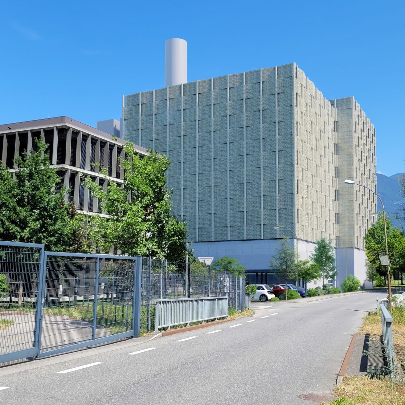 Ein modernes Gebäude mit einem hohen Schornstein steht an einem sonnigen Tag. Bäume und Pflanzen befinden sich in der Nähe. Eine Straße mit geparkten Autos und Straßenlaternen ist in der Nähe.