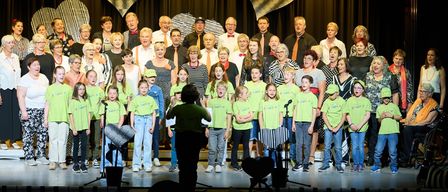 Ein Chor aus verschiedenen Personen, Erwachsene und Kinder, in grünen T-Shirts, stehen mit Mikrofonen auf der Bühne. Sie scheinen zu singen. Dahinter ein schwarzer Vorhang mit Herzdesigns und Scheinwerfern.