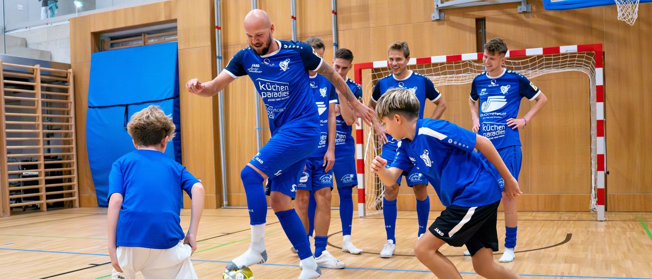Eine Gruppe junger Jungen und Männer spielt Fußball auf einem Spielfeld mit Holzbelag. Ein Junge in einem blauen Trikot tritt den Ball.