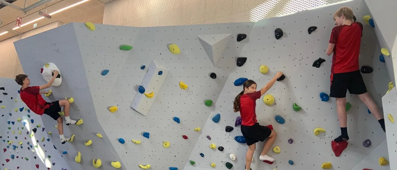 Drei Kinder klettern an einer Kletterwand mit farbigen Griffen, zwei in roten Shirts und schwarzen Shorts, eines mit weißen Socken. Die Wand ist weiß mit blauer Polsterung darunter.