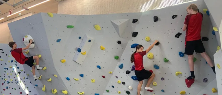 Drei Kinder klettern an einer Kletterwand mit farbigen Griffen, zwei in roten Shirts und schwarzen Shorts, eines mit weißen Socken. Die Wand ist weiß mit blauer Polsterung darunter.