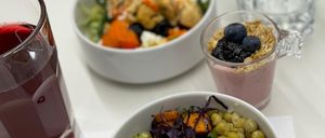 Drei Schüsseln mit Essen auf einem weißen Tisch. Eine Schüssel enthält einen Gemüsesalat mit Brokkoli, Quinoa und Kichererbsen. Eine weitere Schüssel hat einen Smoothie mit Beeren und Granola. Ein Glas Saft ist ebenfalls vorhanden.