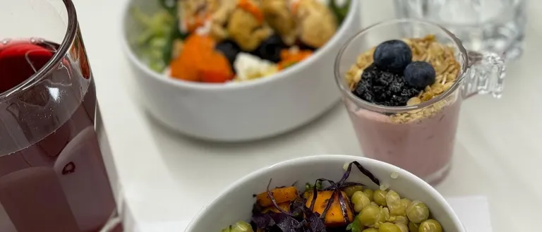 Drei Schüsseln mit Essen auf einem weißen Tisch. Eine Schüssel enthält einen Gemüsesalat mit Brokkoli, Quinoa und Kichererbsen. Eine weitere Schüssel hat einen Smoothie mit Beeren und Granola. Ein Glas Saft ist ebenfalls vorhanden.