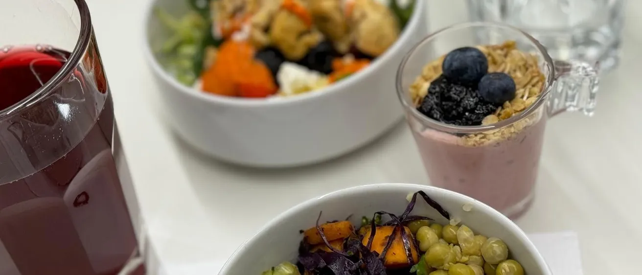 Drei Schüsseln mit Essen auf einem weißen Tisch. Eine Schüssel enthält einen Gemüsesalat mit Brokkoli, Quinoa und Kichererbsen. Eine weitere Schüssel hat einen Smoothie mit Beeren und Granola. Ein Glas Saft ist ebenfalls vorhanden.