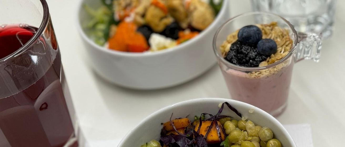 Drei Schüsseln mit Essen auf einem weißen Tisch. Eine Schüssel enthält einen Gemüsesalat mit Brokkoli, Quinoa und Kichererbsen. Eine weitere Schüssel hat einen Smoothie mit Beeren und Granola. Ein Glas Saft ist ebenfalls vorhanden.