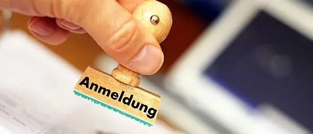 Ein Holzstempel mit dem Wort 'Anmeldung' darauf wird von einer Hand auf ein Stück Papier gedrückt.