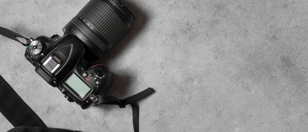 Eine schwarze DSLR-Kamera mit Zoomobjektiv liegt auf einer grauen strukturierten Oberfläche. Das Objektiv der Kamera hat Entfernungsmarkierungen und der Bildschirm der Kamera ist ausgeschaltet.