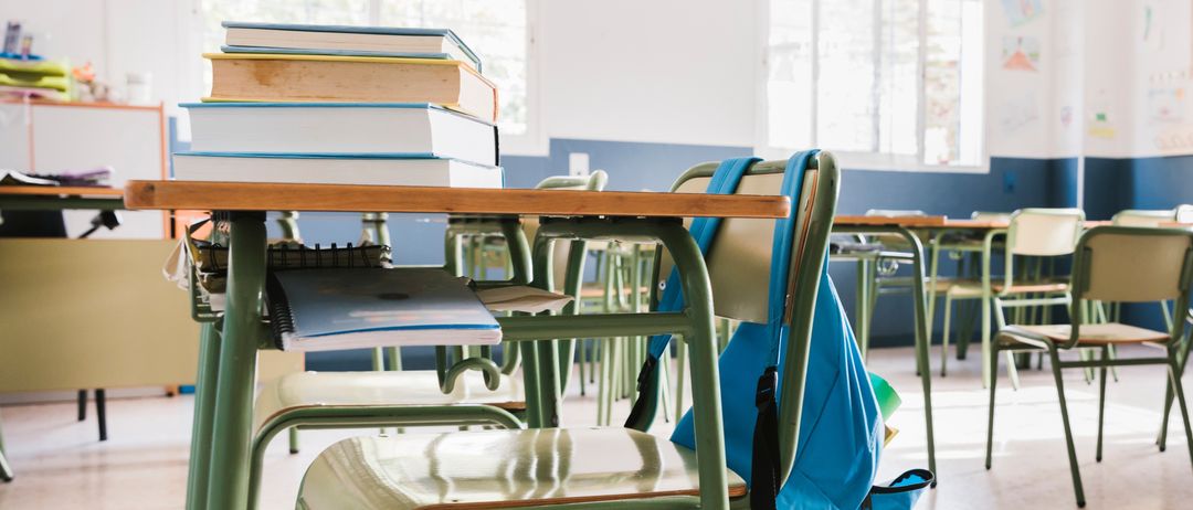Ein leerer Klassenraum mit einem Schreibtisch, Büchern und einem Rucksack. Auf dem Schreibtisch liegen mehrere Bücher. Der Klassenraum hat weiße Wände und blaue Ränder.