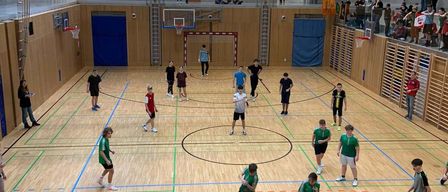 Hallensportanlage mit mehreren Personen, die ein Spiel spielen, einige in grünen Trikots und andere in Schwarz. Der Platz ist mit verschiedenen Linien markiert. Zuschauer beobachten von den Seiten.