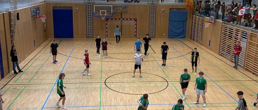 Hallensportanlage mit mehreren Personen, die ein Spiel spielen, einige in grünen Trikots und andere in Schwarz. Der Platz ist mit verschiedenen Linien markiert. Zuschauer beobachten von den Seiten.