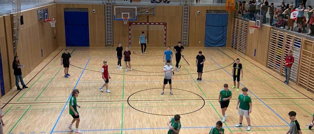 Hallensportanlage mit mehreren Personen, die ein Spiel spielen, einige in grünen Trikots und andere in Schwarz. Der Platz ist mit verschiedenen Linien markiert. Zuschauer beobachten von den Seiten.