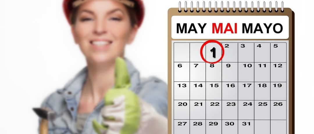 Eine Frau in einem blauen Hemd und einem roten Helm hält einen grünen Handschuh und lächelt. Sie steht neben einem Kalender, der den 1. Mai hervorhebt.