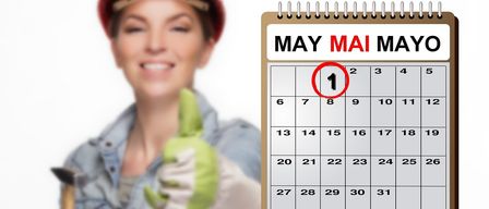 Eine Frau in einem blauen Hemd und einem roten Helm hält einen grünen Handschuh und lächelt. Sie steht neben einem Kalender, der den 1. Mai hervorhebt.