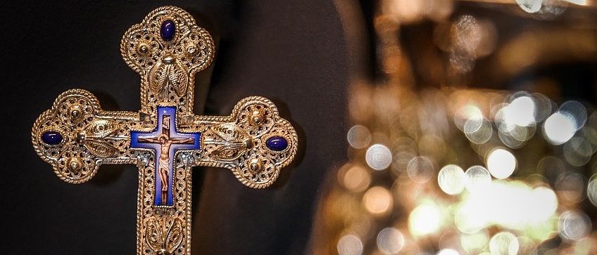 Ein goldenes Kreuz mit aufwendigen Verzierungen und blauen Steinen steht auf einem dunklen Hintergrund neben einem goldenen Kelch mit funkelnden Lichtreflexionen.