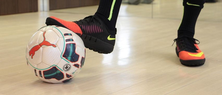 Eine Person tritt einen Fußball mit einem Schuh in einem Fitnessstudio. Der Schuh ist schwarz mit orangenen und pinken Details. Es gibt einen Spiegel an der Wand dahinter.