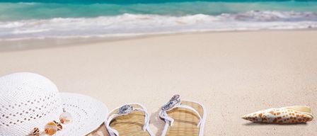 Zwei weiße Flip-Flops mit einem Diamanten auf dem Riemen und ein weißer Hut liegen am Strand. Das Meer ist blau und ruhig, und der Himmel ist blau mit einigen Wolken.