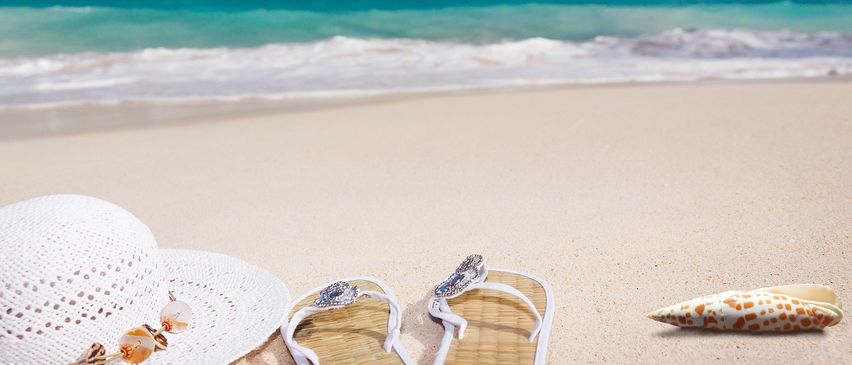Zwei weiße Flip-Flops mit einem Diamanten auf dem Riemen und ein weißer Hut liegen am Strand. Das Meer ist blau und ruhig, und der Himmel ist blau mit einigen Wolken.