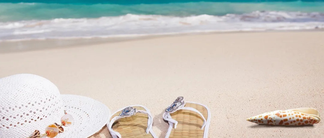 Zwei weiße Flip-Flops mit einem Diamanten auf dem Riemen und ein weißer Hut liegen am Strand. Das Meer ist blau und ruhig, und der Himmel ist blau mit einigen Wolken.