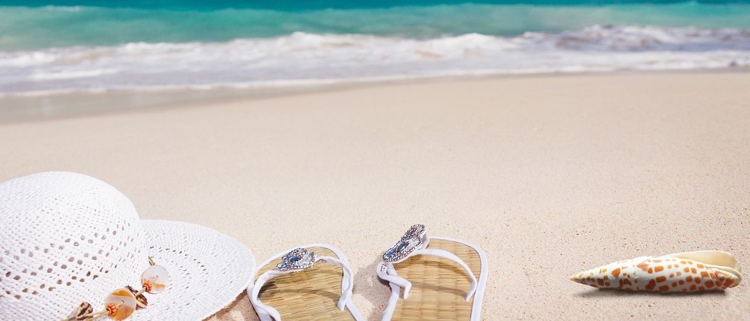 Zwei weiße Flip-Flops mit einem Diamanten auf dem Riemen und ein weißer Hut liegen am Strand. Das Meer ist blau und ruhig, und der Himmel ist blau mit einigen Wolken.