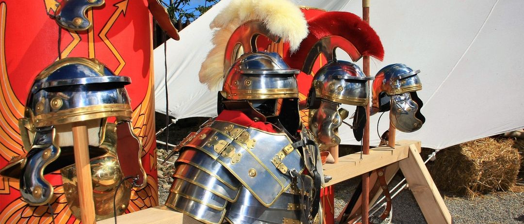 Eine Sammlung antiker römischer Soldatenhelme und Rüstungen, die in einem historischen Umfeld ausgestellt sind, mit einem weißen Zelt im Hintergrund.