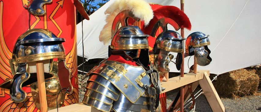 Eine Sammlung antiker römischer Soldatenhelme und Rüstungen, die in einem historischen Umfeld ausgestellt sind, mit einem weißen Zelt im Hintergrund.