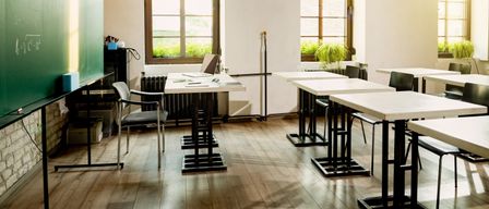 Ein leeres Klassenzimmer mit Holzböden, einer Tafel und weißen Tischen und Stühlen. Es gibt Fenster mit Pflanzen draußen, und eine Heizung ist an der Wand.