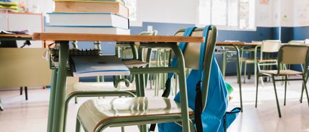 Ein Klassenzimmer mit grünen Tischen, leeren Stühlen und einem blauen Rucksack auf einem der Stühle. Es gibt Stapel von Büchern auf einem Tisch und mehrere leere Tische im Hintergrund.