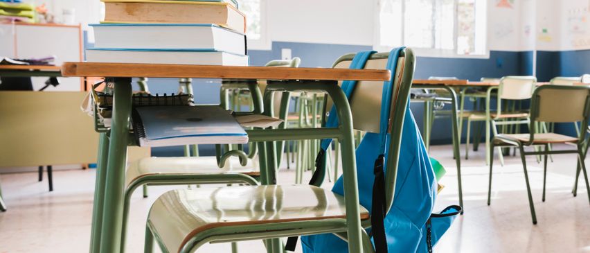 Ein Klassenzimmer mit grünen Tischen, leeren Stühlen und einem blauen Rucksack auf einem der Stühle. Es gibt Stapel von Büchern auf einem Tisch und mehrere leere Tische im Hintergrund.