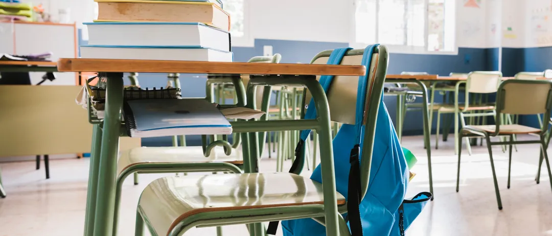 Ein Klassenzimmer mit grünen Tischen, leeren Stühlen und einem blauen Rucksack auf einem der Stühle. Es gibt Stapel von Büchern auf einem Tisch und mehrere leere Tische im Hintergrund.