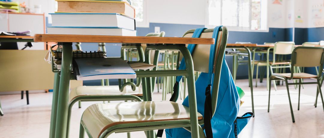 Ein Klassenzimmer mit grünen Tischen, leeren Stühlen und einem blauen Rucksack auf einem der Stühle. Es gibt Stapel von Büchern auf einem Tisch und mehrere leere Tische im Hintergrund.