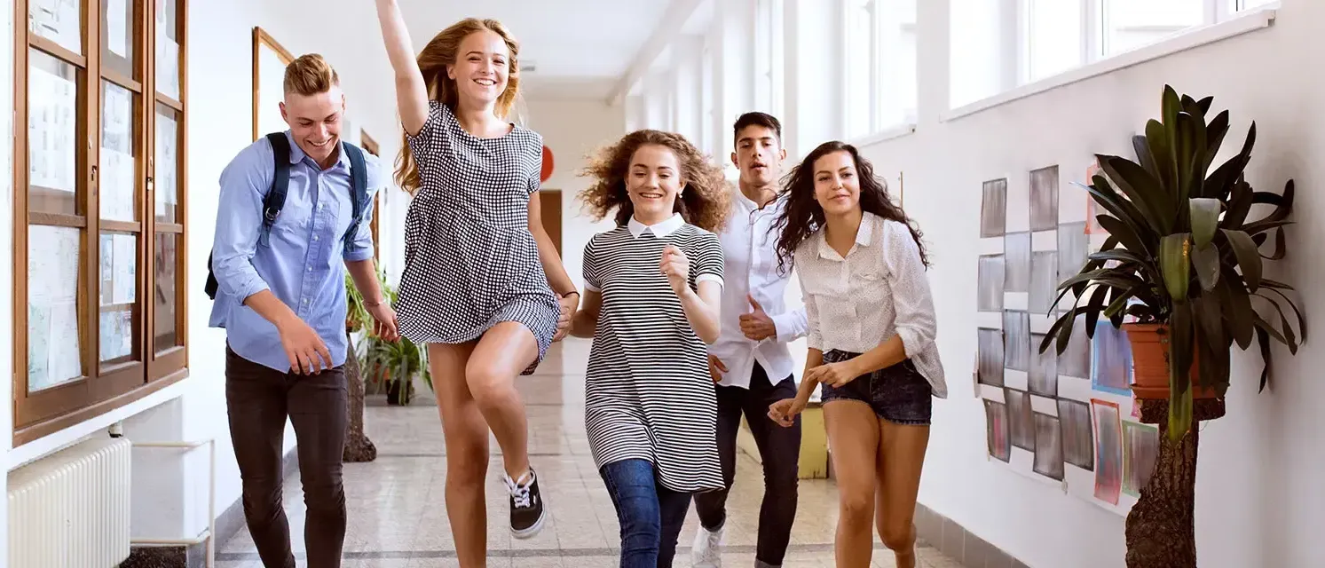 Eine Gruppe junger Erwachsener läuft und lacht in einem Flur, wobei ein Mädchen springt und lächelt und andere sie beobachten.