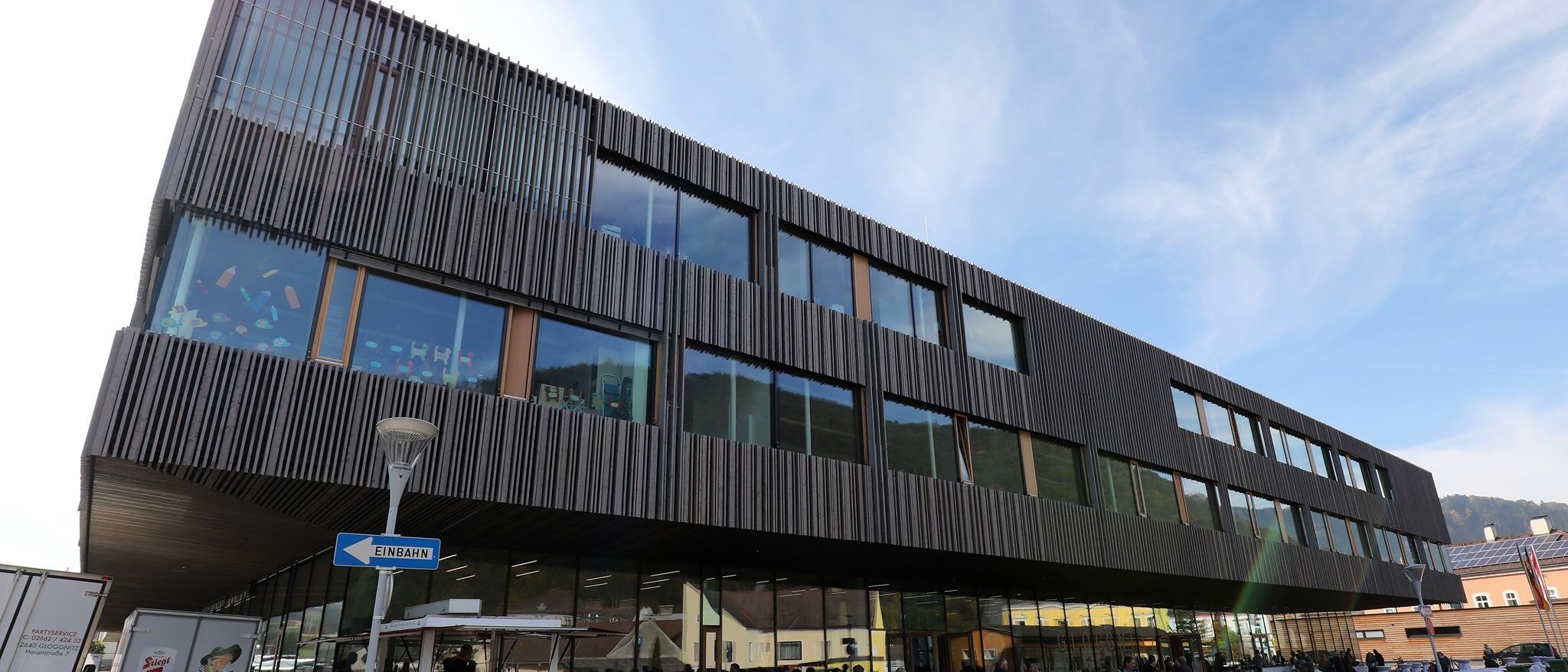 Ein modernes Gebäude mit Glasfenstern und einem Schild für Einbahn. Menschen sitzen draußen an Tischen unter einem blauen Himmel.