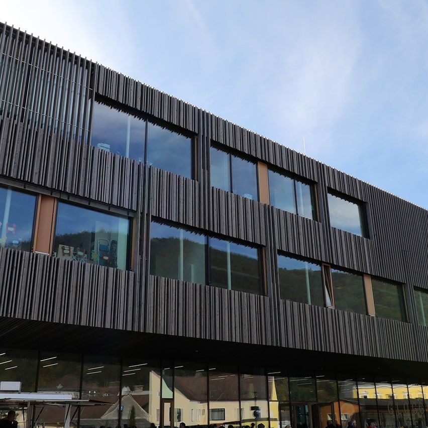 Ein modernes Gebäude mit Glasfenstern und einem Schild für Einbahn. Menschen sitzen draußen an Tischen unter einem blauen Himmel.