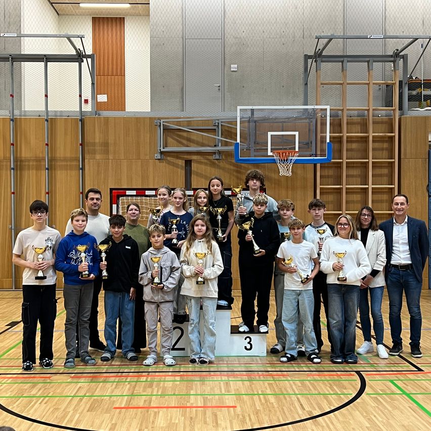 Eine Gruppe junger Athleten und ihre Trainer posieren für ein Foto auf einem Basketballplatz, halten Trophäen und stehen auf einem Podest.