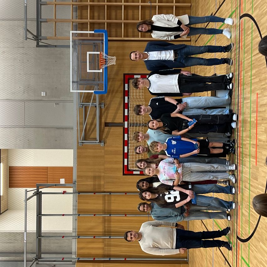 Eine Gruppe von Menschen steht auf einem Basketballplatz, wobei zwei Personen vor ihnen Telefone halten. Hinter ihnen ist ein Basketballkorb aufgestellt. Rechts befindet sich ein Tornetz, neben dem eine Person steht. Der Holzboden hat grüne Markierungen.