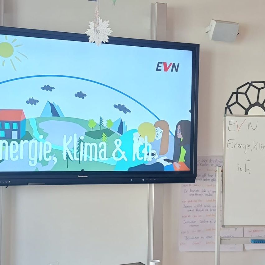 Ein Klassenzimmer mit einem großen Monitor, der ein Bild von einem Haus, Bergen und Menschen mit den Worten 'Energie, Klima & Ich' zeigt. Eine Tafel mit Text steht rechts, und eine Schneeflockendekoration ist an der Wand.