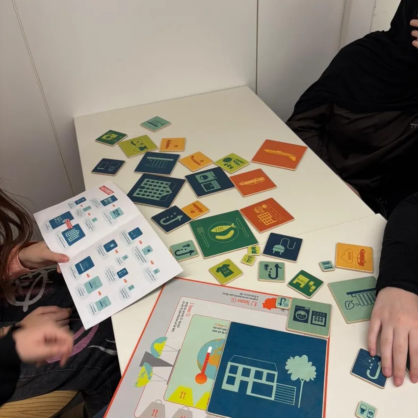 Zwei Personen spielen ein Brettspiel auf einem weißen Tisch mit verschiedenen Karten und Kacheln. Ein Spieler hält ein Büchlein und eine Karte, während der andere mit einem Spielbrett spielt, das Häuser, einen Baum und andere Symbole zeigt.