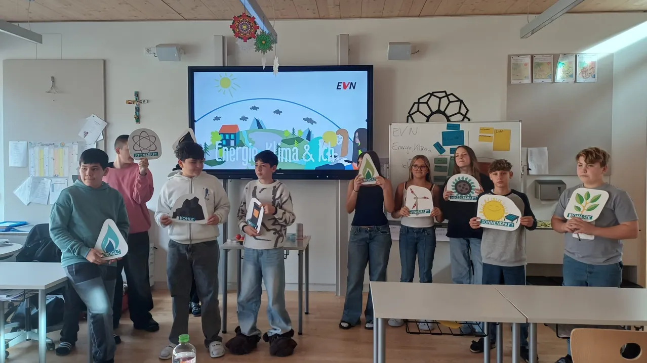 Schüler halten Papierbasteleien hoch, möglicherweise im Zusammenhang mit einem Energie- und Klimaprojekt, vor einem großen Bildschirm, der eine animierte Umweltszene zeigt.