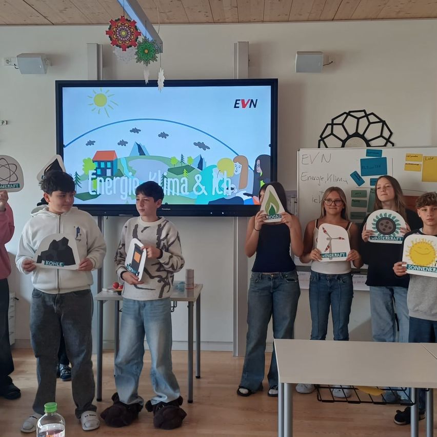 Schüler halten Papierbasteleien hoch, möglicherweise im Zusammenhang mit einem Energie- und Klimaprojekt, vor einem großen Bildschirm, der eine animierte Umweltszene zeigt.