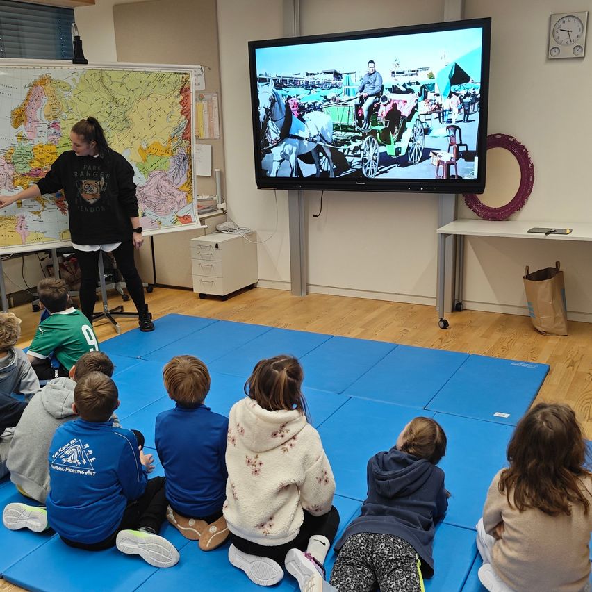 Eine Frau unterrichtet eine Gruppe von Kindern, die auf blauen Matten in einem Klassenzimmer sitzen. Ein großer Bildschirm zeigt einen Mann auf einer Pferdekutsche.