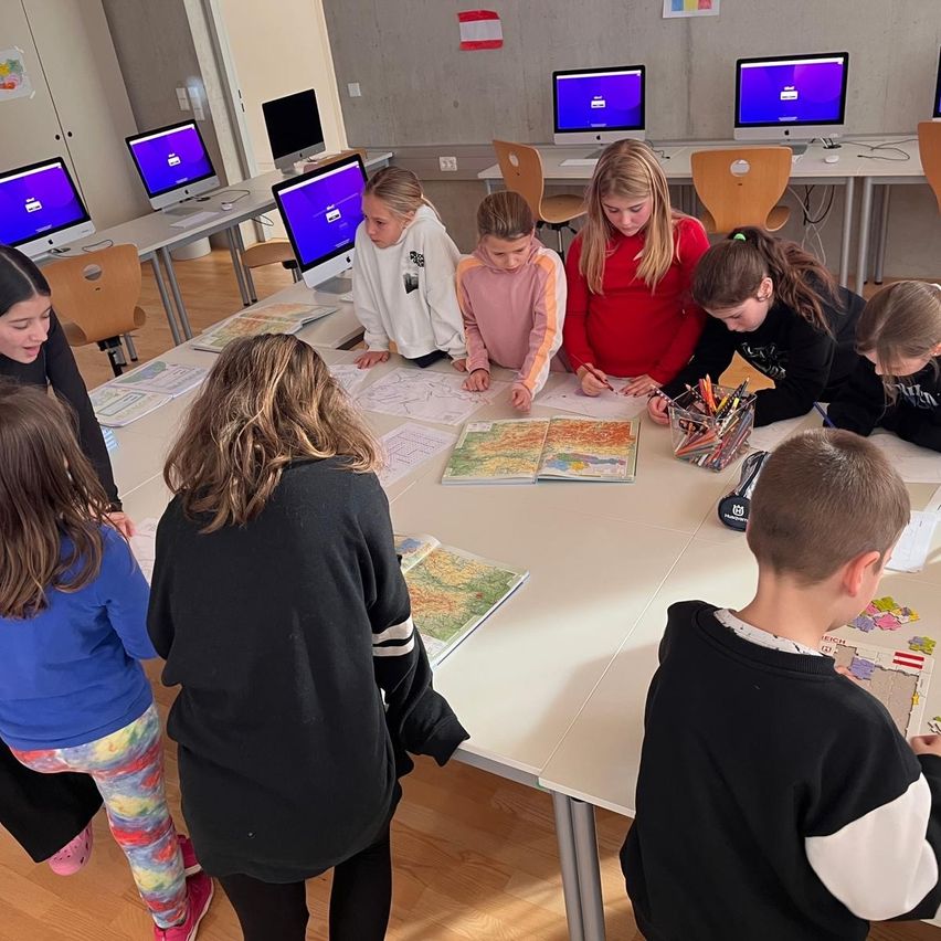 Ein Klassenzimmer mit Schülern an Schreibtischen, einige benutzen Computer. Andere stehen um einen Tisch mit Karten und Malutensilien.