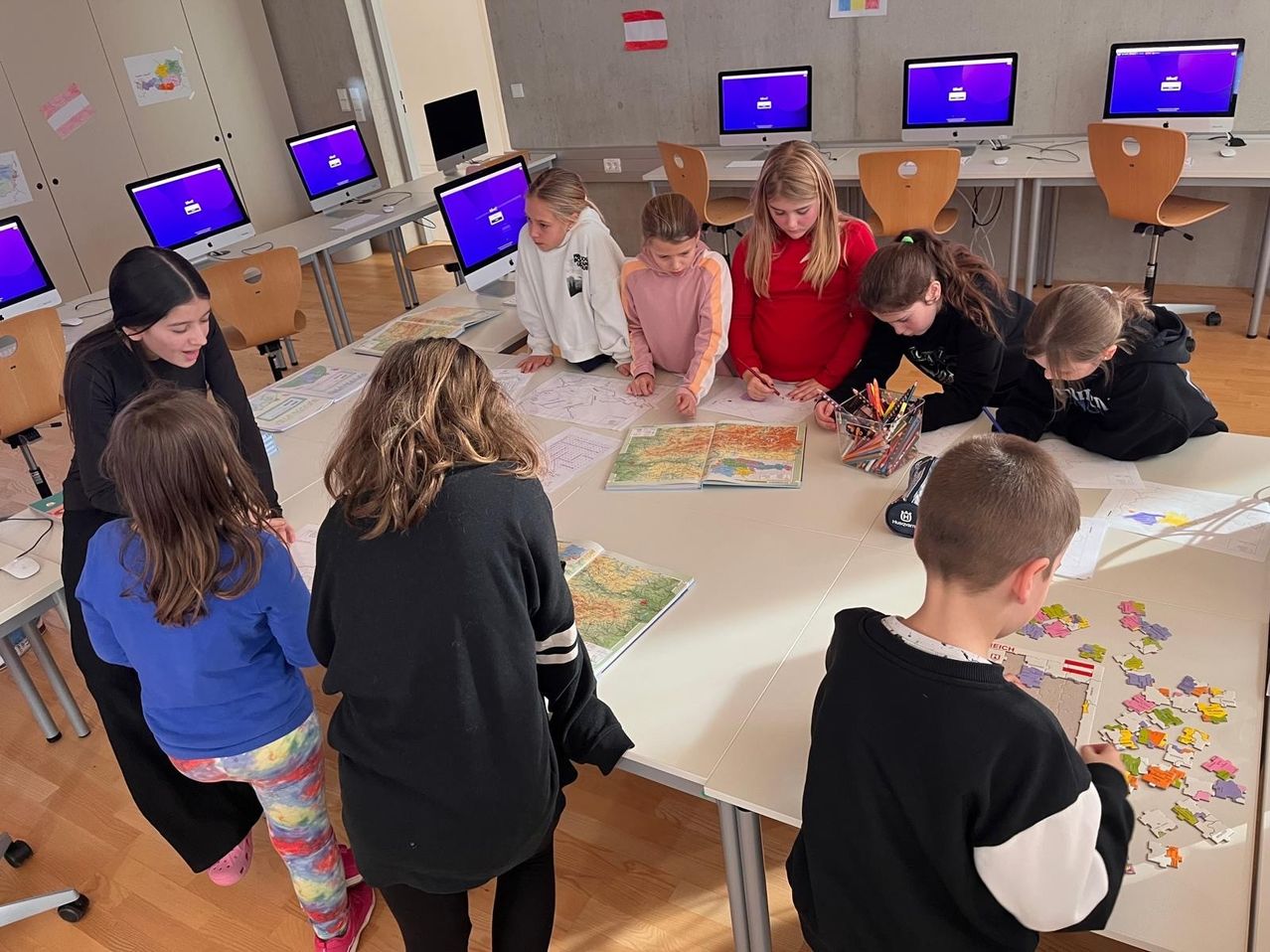 Ein Klassenzimmer mit Schülern an Schreibtischen, einige benutzen Computer. Andere stehen um einen Tisch mit Karten und Malutensilien.