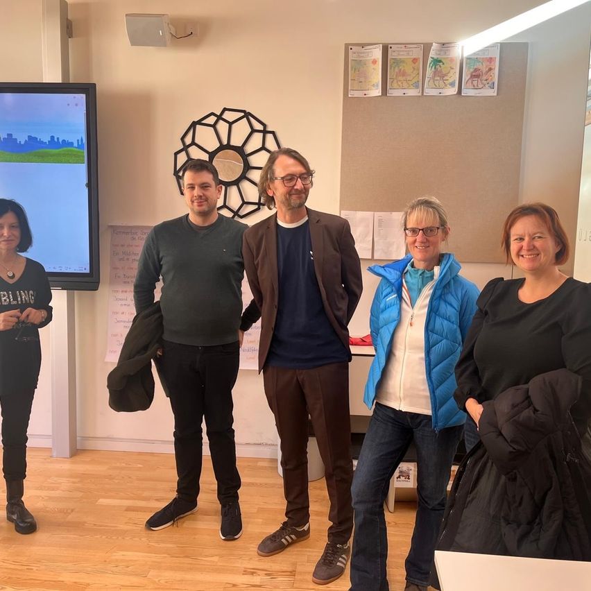 Fünf Erwachsene stehen in einem Raum mit einem großen Bildschirm an der Wand und einer dekorativen Wandbehang. Sie alle lächeln für ein Foto.