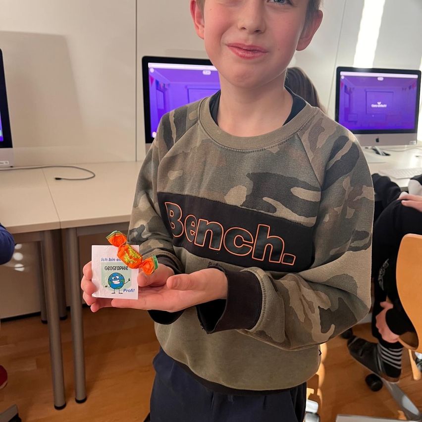 Ein Junge mit einem Tarnsweatshirt hält eine Süßigkeit und eine kleine weiße Karte in den Händen. Er steht in einem Raum mit einem Schreibtisch und Monitoren hinter ihm.