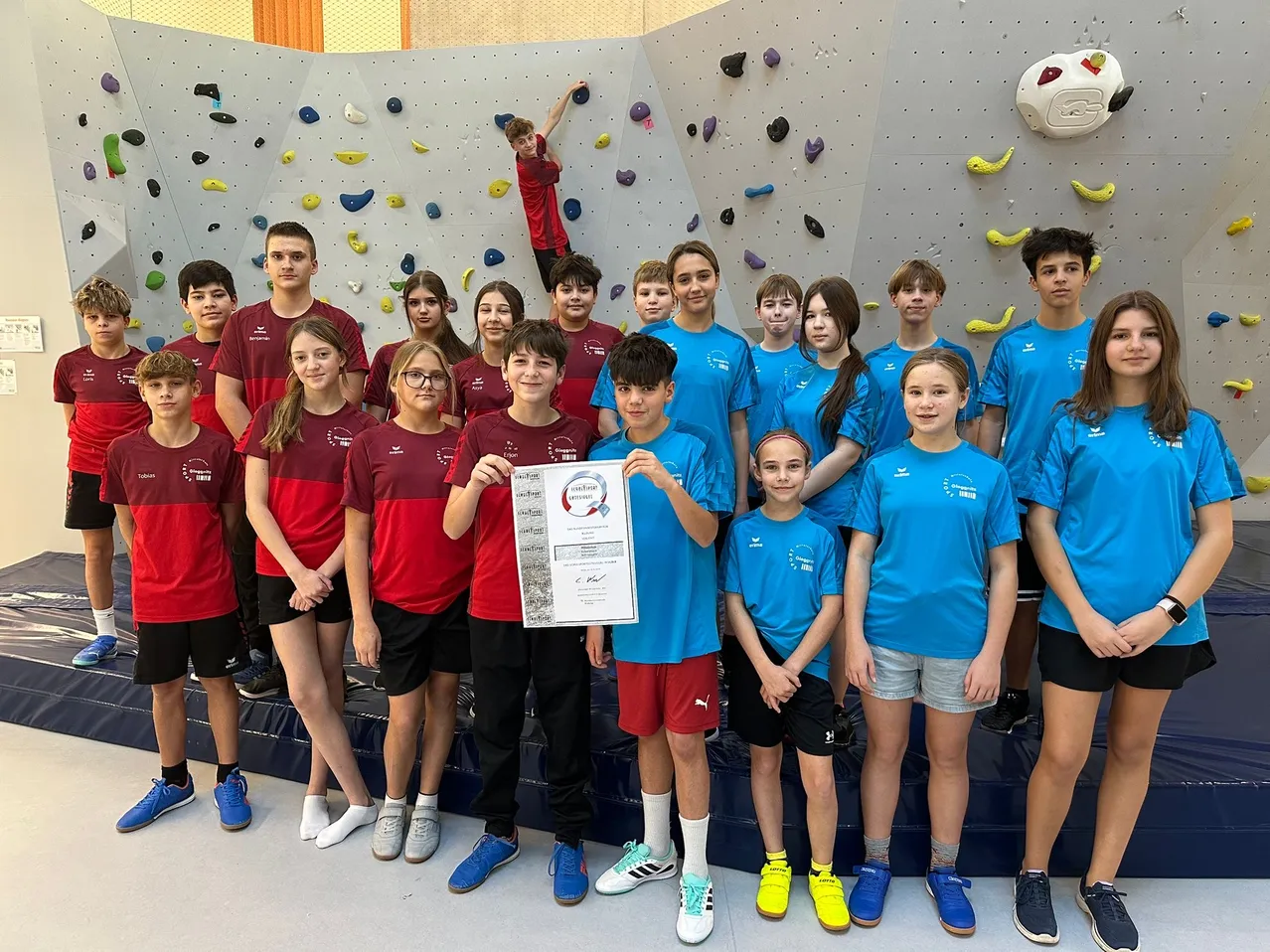 Eine Gruppe junger Athleten in blauen und roten Uniformen posiert für ein Foto vor einer Kletterwand. Ein Junge in der Mitte hält ein Zertifikat.