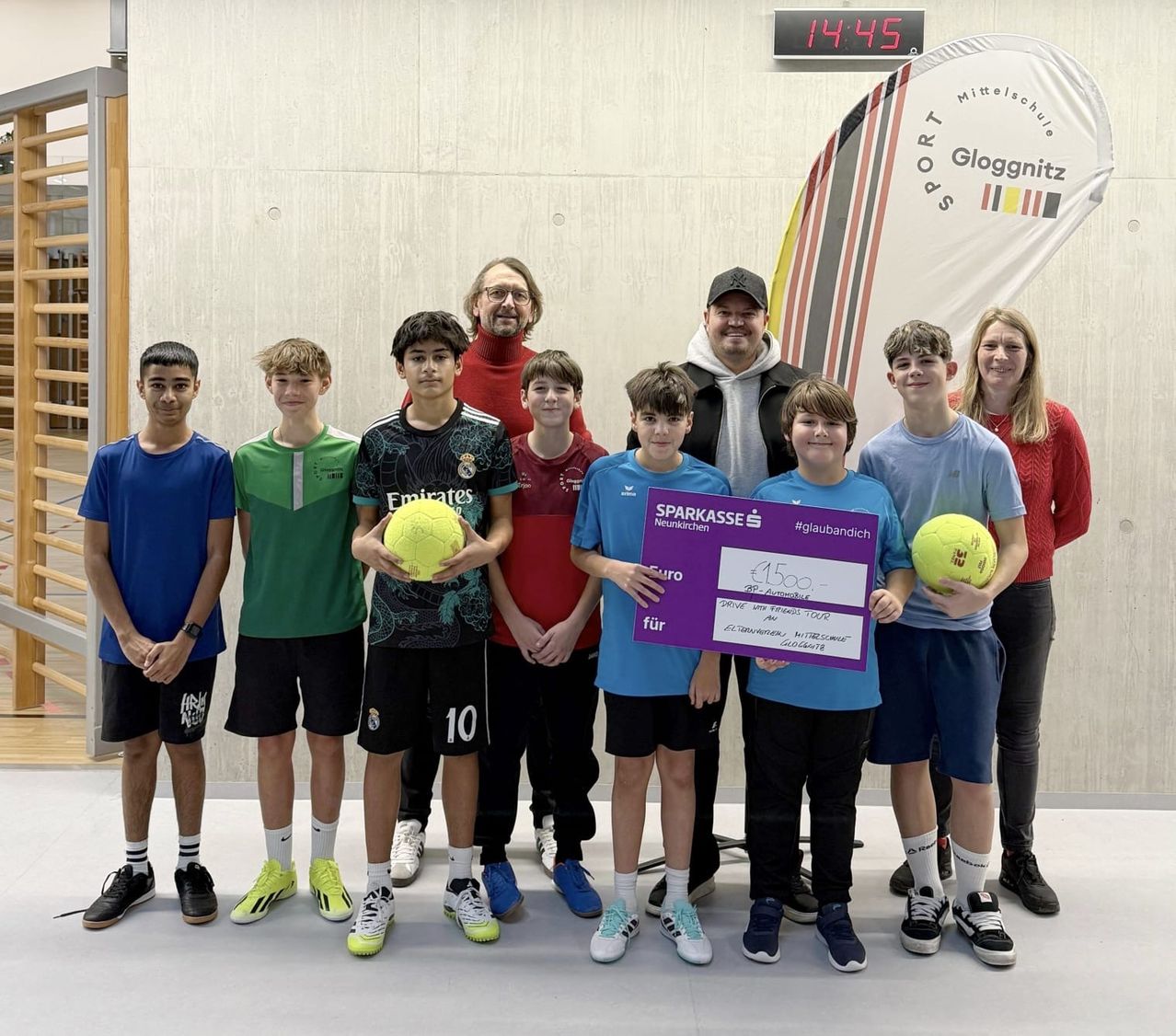 Gruppe junger Fußballspieler und zwei Erwachsene posieren für ein Foto. Sie halten Fußballbälle, einen Scheck und stehen vor einem Banner mit dem Text Gloggnitz.