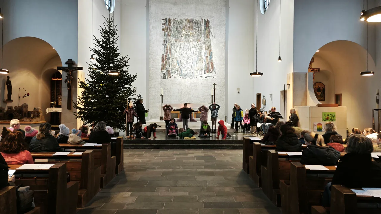 In einer Kirche versammeln sich Kinder und Erwachsene für ein festliches Ereignis. Einige sitzen auf dem Boden, andere stehen in der Nähe eines Weihnachtsbaums. Ein Wandbild schmückt die Wand hinter dem Altar.
