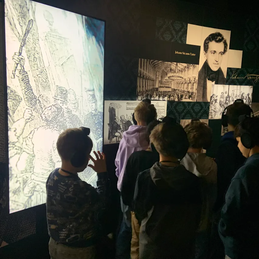Eine Gruppe von Kindern in einem Museum schaut eine große Bildschirmprojektion an, trägt Kopfhörer, und ein Mann zeigt auf den Bildschirm.