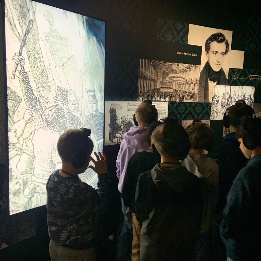 Eine Gruppe von Kindern in einem Museum schaut eine große Bildschirmprojektion an, trägt Kopfhörer, und ein Mann zeigt auf den Bildschirm.